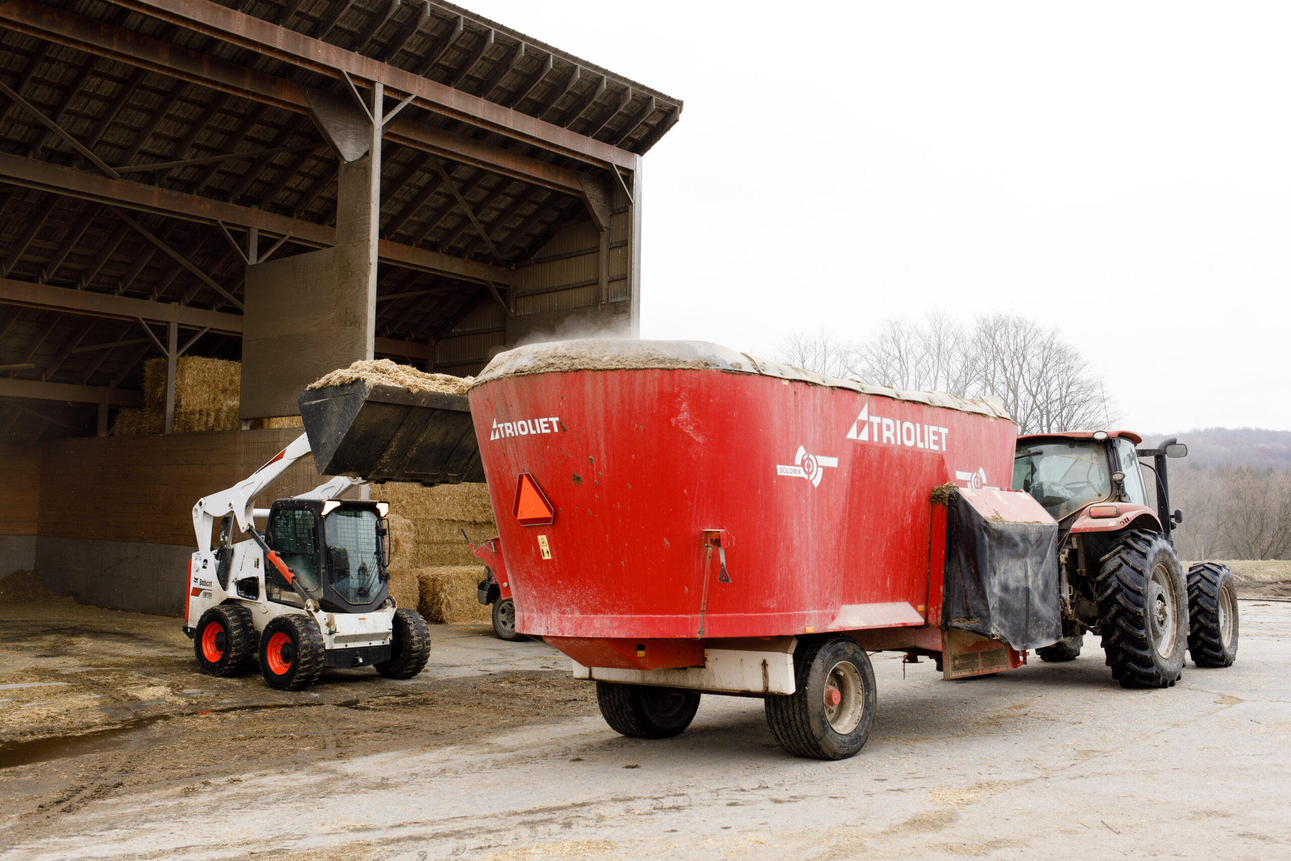 What Do Dairy Cows Eat Blog 1 A machine loads TMR feed, for dairy cows, into a red trailer being pulled by a tractor next to a food shed.